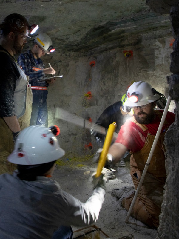 Students on knees working together handing things to each other to wire up a wall rigged with explosives inside a mine other students observe them and other unseen students all student are wearing hard hats with headlamps turned on